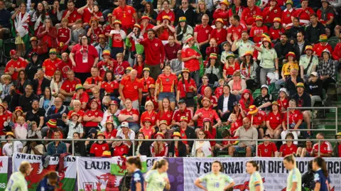 FAW Wales fans in the stands watch Wales against France in St Gallen. Welsh national flags are draped over barriers with Wales players in lime green change shirts stand on the pitch.