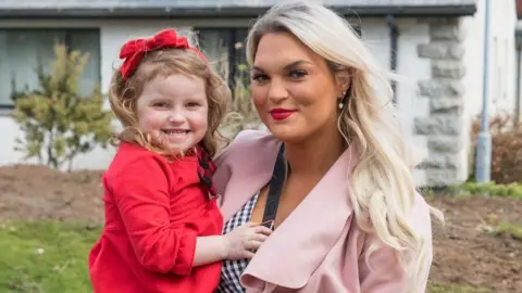 Inishowen Independent Natalie McLaughlin holds her young daughter Ella in her arms. She is smiling, has long blonde hair, and is wearing silver earrings, a pink overcoat and a black and white checked blouse. Ella is also smiling, and has a red bow in her ong brown hair. The little girl is wearing a red dress. Behind them is a garden and house