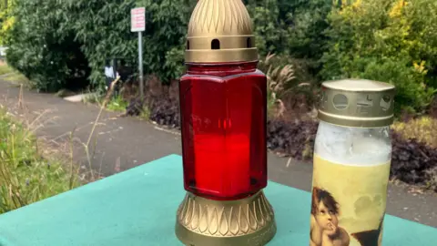 BBC A close-up of two candles left at the scene of a man's murder in Fisher Park, Newry city. One of the candles is in a red and gold glass lantern and the other is in a clear glass and gold jar and is decorated with a cherub-like figure. A shrub-lined path in the park is partially visible behind the candles.