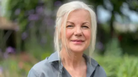 Caroline Eshghi A woman is standing in a garden smiling at the camera. She has grey/silver hair and is wearing a grey shirt.