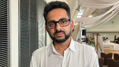 Miten Patel, who is wearing a white shirt and looking into the camera. In the background is a large room decorated with white fabric as part of a memorial event in August.