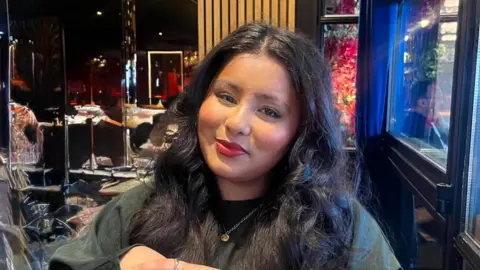 A photo of Hannah Begum sitting in a restaurant. She has long black hair and is wearing a black jumper with a golden pendant. 