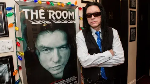 Getty Images Tommy Wiseau with long black hair, black glasses and a black waistcoat leans against a poster for The Room, framed by coloured fairy lights, at a screening in New Orleans in 2014 
