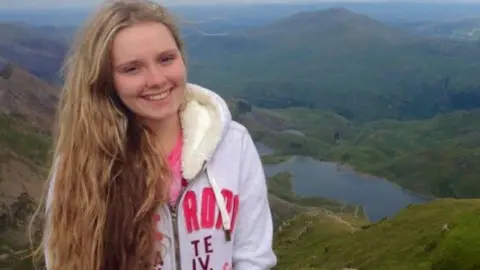 Family A teenage girl smiling at the camera, with long blonde hair, wearing a fur lined hoodie. She is standing high on the side of a mountain with other mountains, trees and a lake in the background.