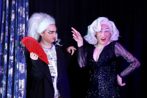 Deaf drag queens Mary and Danielle. Danielle is in a platinum blonde pony tail wig and a black-and-white newspaper print outfit, and holds a red fan while looking at Mary. Mary wears a Marilyn Monroe-style bouffant blonde hairstyle, with a sparkly black dress and has an animated expression on her face.