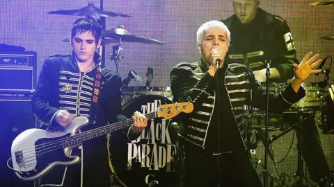 Getty Images Mikey Way and Gerard Way of My Chemical Romance performing onstage at the Scream Awards 2006. Gerard is mid-performance, wearing their signature Black Parade jackets that are black with white fastenings on the front. Former drummer, Bob Bryer, is in the background mid-drum. On the drumset 'The Black Parade' is written in a large font. 