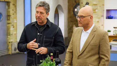 John Torode, left, and Gregg Wallace standing in the studio kitchen. Torode is wearing a black shirt with white rims. Wallace is wearing a brown or cream coloured jacket.