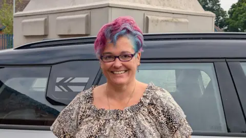 Alison Pope Alison Pope stands against a black car, she is wearing glasses and has a leopard print top. Her hair is dyed blue and pink.  