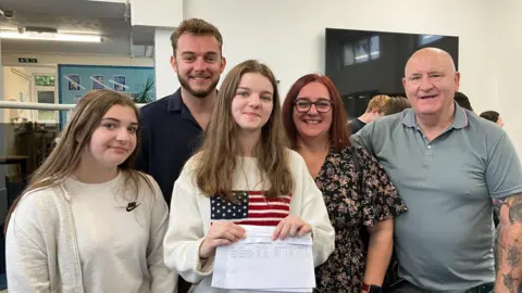 Megan, a young woman with long brown hair wearing a white jumper with the American flag on it, is flanked by her family: A young woman, a young man, and a middle-aged man and woman. They are all smiling at the camera and Megan is holding her results, on a sheet of paper, in front of her.
