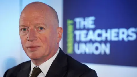 Matt Wrack Matt Wrack is wearing a suit and is sitting in front of a poster which says the teachers union