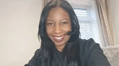 Marie Gentles A woman smiles at the camera. She has long black hair and is wearing a black top, behind her is a cream wall and a window, with cream curtains