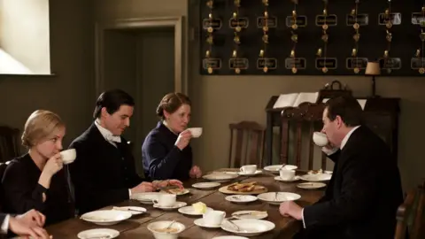 Carnival Films A scene from Downton Abbey. Servants - two men and two women - sit at a large wooden table drinking tea from white cups and saucers. On the wall is a large black board covered in brass butler bells on springs, each labelled with the name of a different room in the house.