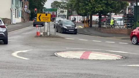 A mini-roundabout painted with the flag of St George - with cars going in two directions and a road sign showing Redditch. 