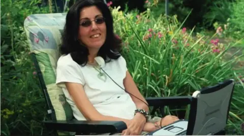 Sarah Ezekiel A family photo of Sarah, sitting in a leafy garden on an upright sun lounger. She is smiling and has her eye gaze technology screen on her lap. She wears a white short-sleeved top, sunglasses and a bracelet on her wrist.