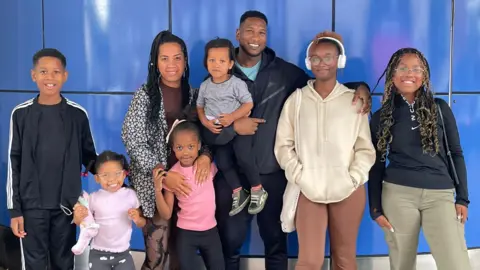 Leon Smith The Smith family of eight stand looking at the camera. They all have their hands around each other and are smiling.