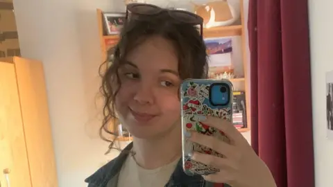 Jess Cooper Jess has brown curly hair tied back. She is smiling into her camera taking a selfie. There is a red curtain and brown wooden door behind her.