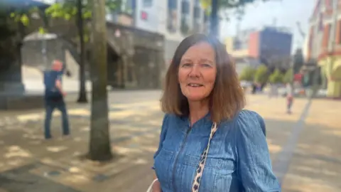 A smiling lady stands in Derry's Guildhall square. The city's walls are seen in the background as are a number of shops. She is wearing a denim top and has long brown hair. A strap of a handbag can be seen over her shoulder