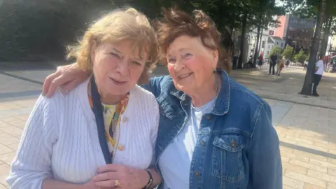 Two ladies stand in Derry city centre in an embrace. The lady on the right is wearing a white cardgian and is holding a balc bag. she has shoulder lenght brown hair . To her right another lady is smiling, she has her arm around her companion. She is wearing a denim jacket and a white tshirt. Both ladies are standing in front of derry's walls/