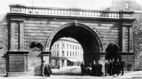 Getty Images A early 20th century black and white photogrpah of Ferryquay Gate, one of the gates of Derry's walls. An archway can be seen in the old ramparts, through which shops can be seen., A number of men stand at the foot of the gate, as the walls reach up above them