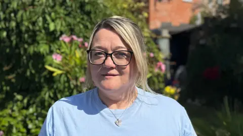 George King/BBC Samantha Mitchell standing in her garden. She is wearing a pale blue T-shirt and glasses. She is looking into the camera.