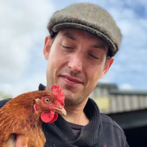 Lewis Clare Lewis is wearing a dark sweater with a tweed flat cap on his head. He holds a gingery-brown chicken close to his chest while standing outside a barn, with a partly cloudy sky and part of a building visible in the background.