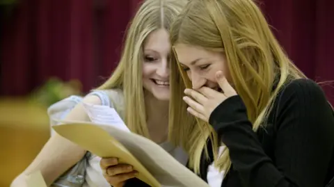 Getty Images Two girls with long blonde hair are smiling while looking at a piece of paper with exam results 