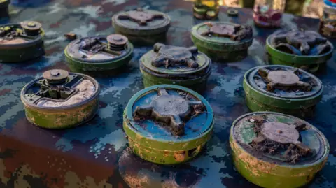 BBC/ Jonathan Head Ten landmines are laid out on a table covered with a camoflauge print cloth. The mines are green and have a cross pattern atop them. Some have soil on them, indicating that they have been recently dug up.