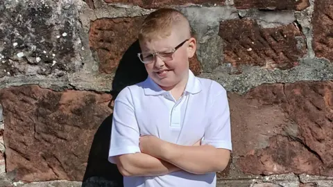 A photo of a 10-year-old boy standing hy a brick wall wearing a white polo shirt. He has short blonde hair and is wearing glasses