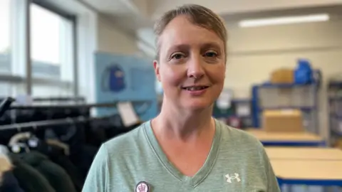 Photo of Julia in the offices of the charity. the background is blurred but we can see storage and school uniform blazers on racks. Julia has short grey hair and is wearing a green v-neck t-shirt
