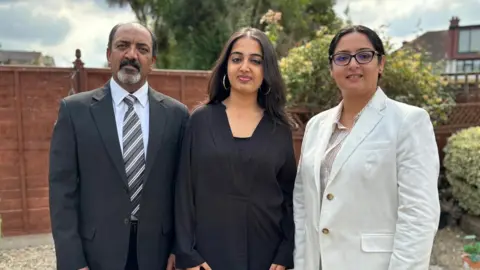 Ms Cheema, a woman with long dark hair and wearing a black top, standing between her parents. Her mother on the right is wearing glasses and a white blazer and has her dark hair tied in a low bun and her father on the left has a greying beard and is wearing a black suit with a stripy tie. They're standing in a garden with a wooden fence behind them. It's a cloudy day.