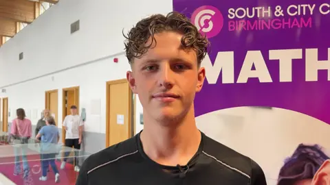 BBC News/Christopher Steers James Bonning, a young man with curly brown hair, stands in a school room in front of a sign saying "South & City College Birmingham". He's wearing a black sports T-shirt and is smiling at the camera.