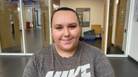 BBC News/Christopher Steers Bethany Clarke, a young woman, is standing in a school hallway. She's wearing a grey Nike T-shirt and has her hair tied back in a ponytail. She is smiling at the camera.