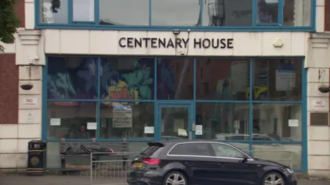 A close up image of the front of Centenary House - the name is spelled out in a metal sign above the entrance which is made up of several plate glass windows and a door. The building is red brick with Portland stone surrounding the entrance. A black Audi is driving past in the foreground.