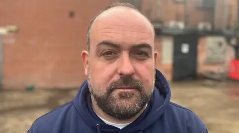 Paul  McCusker is looking directly at the camera. It is a head and shoulders shot. He is wearing a navy hoodie and has short brown hair with a beard. Behind him red brick walls can be seen.