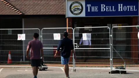 Reuters Two men wearing T-shirts and shorts, one has a rucksack they are walking towards a hotel building covered by a metal barrier. Both have their backs to the camera.