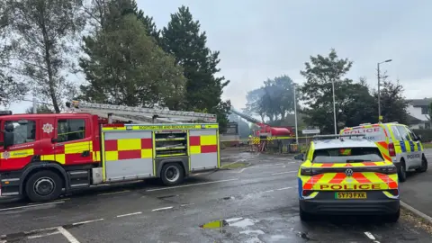Fire engines and police cars parked on a road with trees on a cloudy day