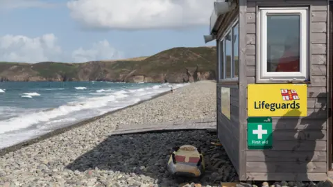 RNLI/Barbara Szente A lifeguard hut on a rocky beach. The hut has a surf board in front of it and a first aid sign on the side. The sea is in the background with waves moving in towards the beach. In the distance are a couple of people on the beach, and beyond them are tall cliffs. The sky is blue with some patchy cloud.