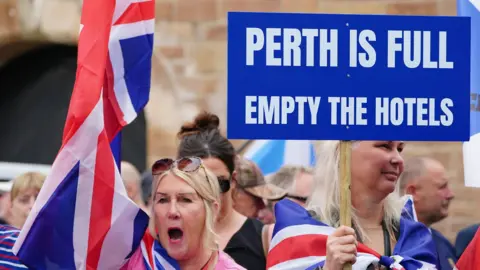 PA Media People demonstrating at an Abolish Asylum System protest outside the Radisson Hotel in Perth. Two blonde women are wrapped in Union Jack flags and one holds a sign which reads "Perth is full empty the hotels"