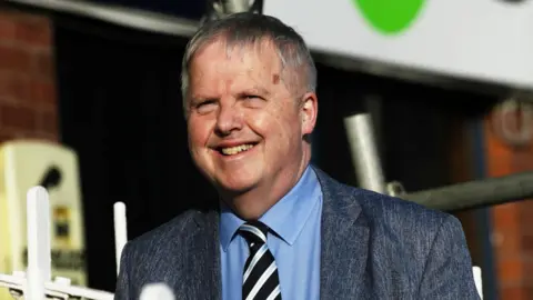 SNS A man with short grey hair smiles. He wears a grey suit with a blue shirt and a striped black and silver tie.