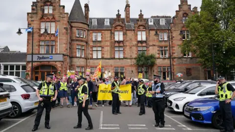 PA Media A group of protestors holding signs which say things like "Perth against racism" stand by the entrance to the Radisson Blu hotel. The entrance leads on to a car park with parked cars. Police stand closer to the camera, in front of the protestors.