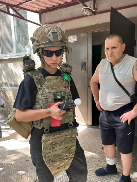 Young woman Varia wearing khaki camouflage gear holding a device, standing next to a middle-aged man wearing a grey vest and blue trousers