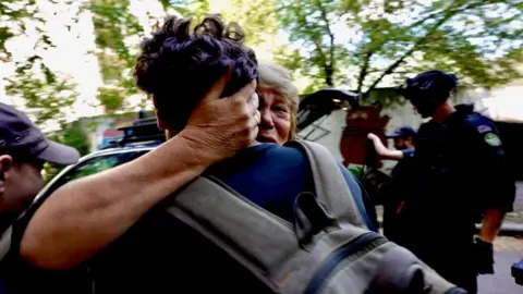 BBC News A blonde woman, who looks distressed, tightly embraces a man with short brown hair. Her hand is round the back of his head and only the back of his head, back and rucksack can be seen.