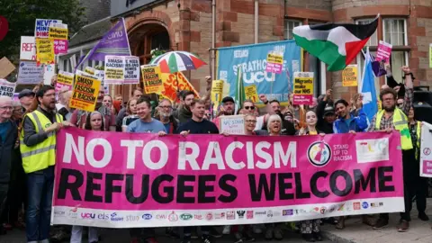 PA Media Dozens of protesters line up behind a large pink "refugees welcome" sign. It is held up by the front row, with others holding signs above their heads. They are standing in front of a stone building.