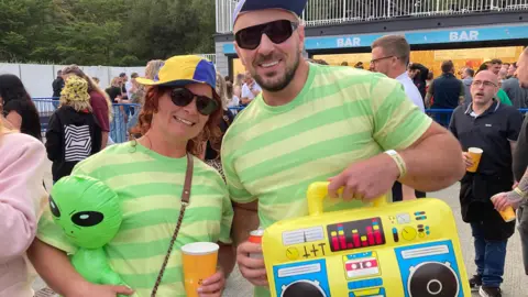 Bek Homer/BBC A woman and a man in green striped T-shirts. The woman is holding a blow-up green alien and the man is holding a blow-up boom box. There are other people milling around in the background.