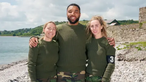 Channel 4 Lucy Spraggan, Troy Deeney and Michaella McCollum stand on a beach with their arms around each other, smiling at the camera. The three are wearing green army style jumpers and camo trousers.