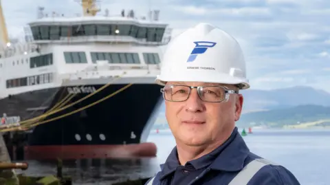 Ferguson Marine A man with a hard hat, wearing glasses, with a large ship in the background 