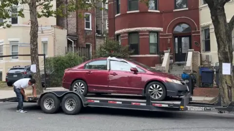 A red car with a smashed window is on a trailer
