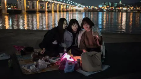Getty Images A group of women smile and look at a mobile phone one of them is holding up  as they pose for a selfie before the Banpo bridge on October 13, 2016. They are sitting on a blanket on a patch of grass, dressed in warm clothes. In front of them are food containers and their bags. Behind them is a lake and the Seoul city skyline.    
