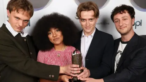 PA Media English Teacher with their trophy for the Mercury Prize. The three men in the band are wearing black jackets, two with white button-up shirts and one with a white T-shirt with black design and collar. Second from the left, the female band member has a voluminous black Afro hairstyle and wears a red and white patterned top.