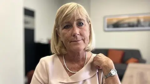 Author of the report, Gladys Rhodes White OBE. She has a blonde bob, with a full fringe and blue eyes. She wears a white pearl necklace and a pale pink top. She holds her right hand up to her shoulder, and on it is a silver watch with a white face. 
